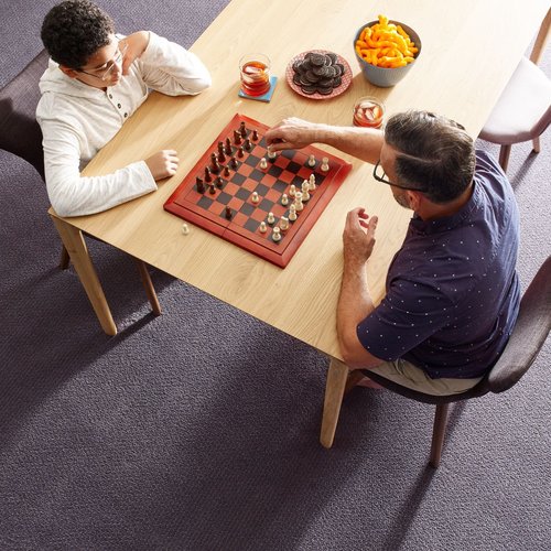 Two people playing chess at a rectangular wooden table in a room with purple carpet from Flooring Source in the Auburn, MA area